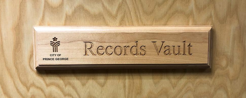 An engraved wooden sign mounted on a wooden door. Sign has a City of Prince George logo. Text reads: Records Vault