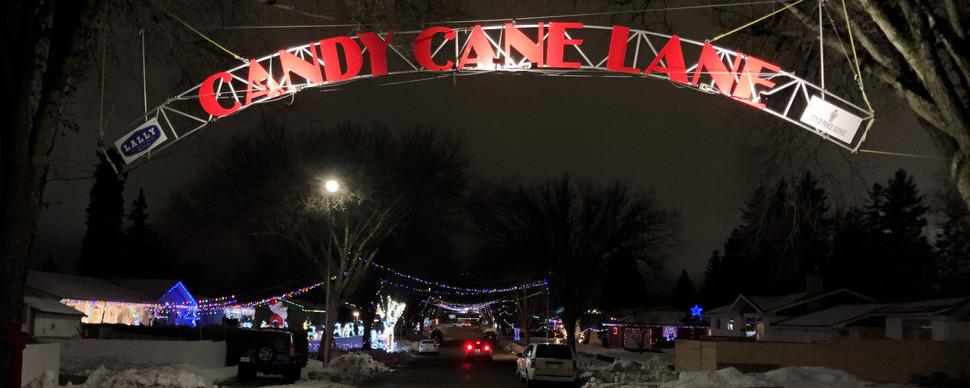 A sign across a roadway that reads Candy Cane Lane. Houses with Christmas lights in the background. Photo taken at night.